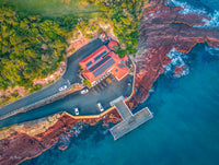 Merimbula Wharf, NSW — aerial view of the red-roofed wharf building and jetty above red rock ledges and turquoise water