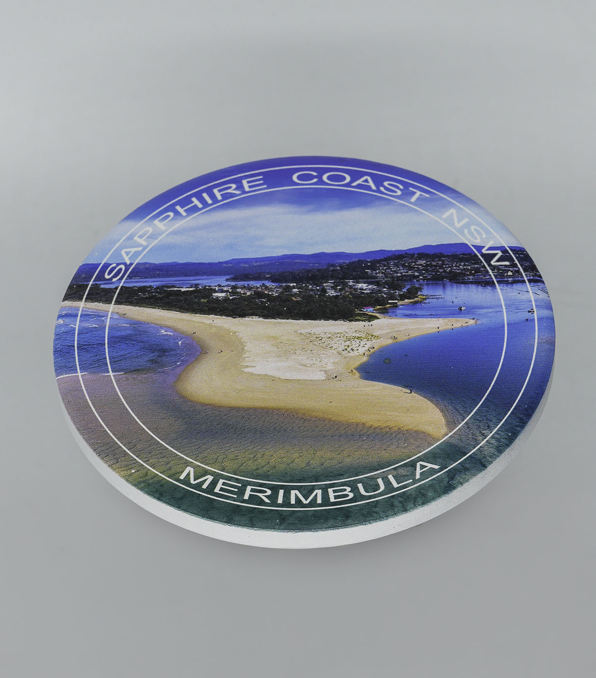 Single round ceramic coaster with location text — Merimbula Lake and Fishpen aerial, NSW